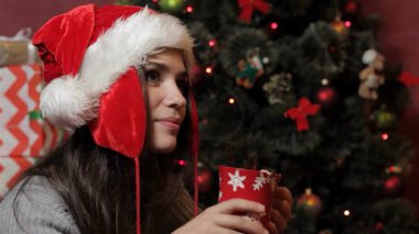 Noel Baba şapkası takmış neşeli, genç, beyaz bir kadın buharlı bir fincan sıcak çikolatanın, sıcaklığın ve bayram neşesinin tadını çıkarıyor. Noel 'de yanıp sönen ışıklar ve renkli süslerle çevrili.