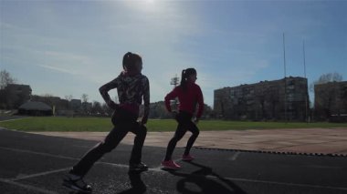 İki genç kız güneşli bir pistte dinamik eğitim egzersizleri yapıyor ve kararlılıklarını ve atletizmlerini sergiliyorlar. Kıyafetlerinin canlı renkleri, açık mavi renge karşı güzel bir tezat oluşturuyor.