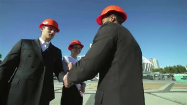 Üç kendine güvenen şık takım elbiseli ve parlak turuncu kasklı mimar, bir inşaat alanında canlı bir tartışmaya girişerek, yenilikçi bina fikirlerini paylaşırken el sıkışıyorlar. Bu canlı sahne.
