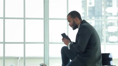 Kendinden emin genç bir iş adamı masasının kenarında oturmuş, akıllı telefonundan gelen aramaları dikkatle kontrol ediyor. Gösterişli bir takım elbise giymiş, profesyonelliği ve kararlılığı parlak bir şekilde temsil ediyor.