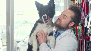 Neşeli, sakallı bir veteriner, neşeli, siyah ve beyaz bir köpeği sevgiyle besliyor. Parlak kliniğinde, sıcaklık ve bakım saçıyor. Yakışıklı erkek veteriner neşeyle ışıldıyor, sağlık kontrolünden sonra mutlu köpekle güçlü bir bağ kuruyor.