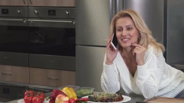 Parlak, olgun, dalgalı sarı saçlı bir kadın elinde canlı bir domates tutarak akıllı telefonuyla sohbet ederken ışıl ışıl gülümsüyor. Bir dizi taze sebze ve yemek zevkleri ile çevrelenmiş, neşeli bir ev yemeğinin özünü somutlaştırıyor.