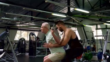 Dinamik bir spor salonu ortamında, odaklanmış genç bir erkek antrenör çömelme ve güvenliği sağlamak için kıdemli bir müşteriye destek olur. İyi bakımlı sakallı kendini işine adamış antrenör yaşlı adamın kollarını tutuyor ve güven duygusu besliyor.