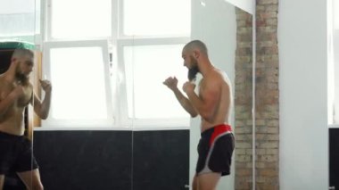 Dinamik bir çekim, iyi aydınlatılmış bir spor salonunda güçlü yumruklar atan üstsüz bir erkek MMA dövüşçüsünü yakalar ve aynadaki kararlılığını yansıtır. Bu sahne gücün, çevikliğin ve acımasız motivasyonun özünü temsil ediyor.