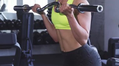 Odaklanmış bir sporcu kadın, modern bir spor salonunda kaslı vücudunu ve kaslarını ustalıkla biceps kıvrımlarını bir ez kıvrım çubuğuyla sergiliyor. Bu dinamik sahne gücün ve adanmışlığın özünü somutlaştırıyor.