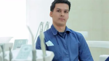 Mavi üniformalı neşeli bir erkek dişçi modern bir diş muayenehanesinde gülümserken başparmak kaldırıyor. Sıcak tavırları ve iyi donanımlı çevresi birinci sınıf diş bakımı isteyen hastalar için hoş karşılayıcı bir ortam yaratıyor.