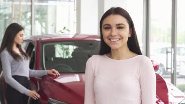 Neşeli genç bir kadın heyecanla ışıldıyor, bir elinde araba anahtarlarını gösteriyor ve yeni arabasının önünde başparmağını kaldırıyor. Satıcının canlı atmosferi, onun mutluluğunu ve başarı duygusunu vurguluyor.