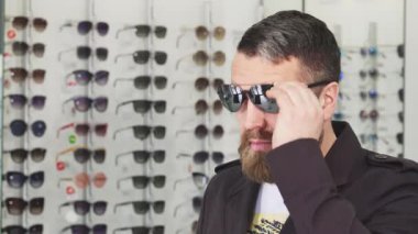 Sakallı, olgun bir adamın canlı bir gözlük mağazasında şık güneş gözlüğü denediği yakın plan bir fotoğraf. Bu yakışıklı müşteri, mükemmel bir aksesuar alırken şık bir yaşam tarzını somutlaştıran çeşitli moda çerçeveleri keşfediyor.