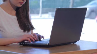 Güzel, beyaz bir kızın laptopuna dikkatlice yazı yazarken, büyüleyici bir kafede işine dalmış bir fotoğrafı. Pencereden süzülen yumuşak ışık, onun rahat beyaz tişörtünü ve davetkar atmosferini ön plana çıkarıyor.