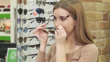 Uzun saçlı neşeli bir kadın optometrist mağazasında son moda güneş gözlüğü deniyor, neşe ve güven saçıyor. Bir dizi modaya uygun göz giyimiyle çevrelenmiş, yaz stili ve kendini ifade etmenin özünü somutlaştırıyor, şık bir seçim yapıyor.