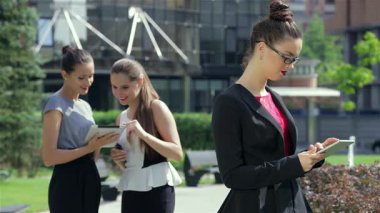 Pürüzsüz siyah takım elbiseli kendine güvenen genç bir iş kadını, açık hava ofis ortamında nişanlısı ile birlikte tabletinin üzerinde çalışıyor. Sahne profesyonelliği ve işbirliğini yayıyor, dinamik enerjiyi gösteriyor