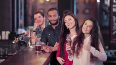 Canlı bir grup genç arkadaş, iki erkek ve iki kadın, hareketli bir barda toplanmış, neşeyle bira bardaklarını kaldırırken başparmaklarını kaldırıyorlar. Bulaşıcı gülümsemeleri ve enerjik enerjileri, neşeli bir atmosfer yaratıyor.