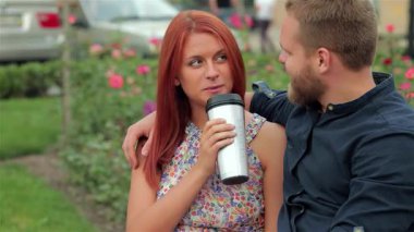 Neşeli bir çift, çiçek açan güllerle çevriliyken şık bir bardaktan kahve yudumlayarak parkta bir banka yakın otururlar. Neşeli gülümsemeleri ve samimi kucaklaşmaları canlı şehir ortamında sıcak bir sevgi ve bağlantı ortamı yaratıyor.