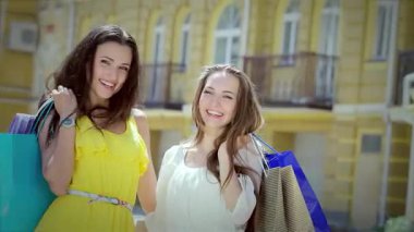 İki neşeli kız, biri parlak sarı elbiseli, diğeri beyaz bluzlu, şık alışveriş çantalarını canlı bir şehir zeminine karşı mutlu bir şekilde sergiliyorlar. Işıl ışıl gülüşleri ve kaygısız ruhları dostluğun özünü yakalar.