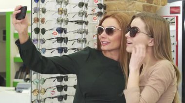 İki şık kadın, biri göz kamaştırıcı kızıl saçlı ve diğeri sarı bukleli, şık bir gözlük mağazasında neşe içinde selfie çekiyor, bir dizi moda güneş gözlüğü ile çevrili. Parlak gülümsemeleri ve neşeli pozları alışverişin heyecanını yansıtıyor.