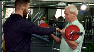 Kararlı, gri saçlı, yaşlı bir adam spor salonunda pazılarını çalıştırmaya odaklanır. Halteri titizlikle kaldırır. Genç Kafkasyalı antrenörü uzman tavsiyesi sunuyor. Bu canlı fitness sahasında destekleyici bir atmosfer yaratıyor.