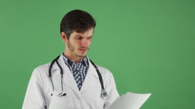 Ciddi ve çekici bir erkek doktor, boynunda steteskop olan çıtır çıtır bir laboratuvar önlüğü giymiş, canlı yeşil renkli bir arka planda tıbbi belgeleri dikkatle inceliyor. Bu görüntü profesyonelliğin ve adanmışlığın özünü yakalar.