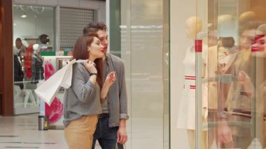 Neşeli genç bir çift, canlı bir mağazanın vitrinindeki şık kıyafetleri inceleyen son moda eğilimlerine hayran kalıyor. Neşeli kadın, neşeli bir gülümsemeyle, erkek arkadaşıyla canlı sohbetler yaparken alışveriş çantalarını tutar,