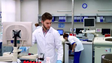 Gıcır gıcır beyaz laboratuvar önlüğü giymiş genç bir beyaz erkek bilim adamı laboratuvar düzeneğini dikkatle inceliyor, eli bir bilgisayar ekranının yanında duruyor. Modern labaratuvar, ileri teknoloji ve canlı renklerle dolup taşıyor.