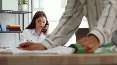 Odaklanmış genç bir bayan moda tasarımcısı, renkli kumaşlar ve sanatının araçlarıyla çevrili bir telefon konuşmasında gülümsüyor. Ön planda, bir meslektaşım titizlikle kumaşı bir mezura ile ölçüyor.