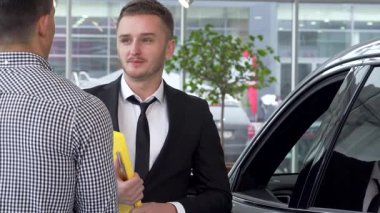 Keskin takım elbiseli karizmatik bir araba satıcısı, şık bir otomobilin başarılı satışını kutlayan tatmin olmuş bir erkek müşteriyle el sıkışıyor. Parlak bayilik ortamı heyecan dolu, modern araçlar sergileniyor ve hoş geldin diyor.