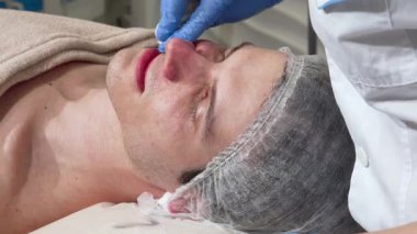 Yetenekli bir kozmetikçi pamuk pedi kullanarak rahat bir erkek müşterinin derisini nazikçe temizler ve sakin bir spa ortamı sunar. Bu samimi an, huzurlu bir ortamda saflık ve gençleşmeyi vurgulayan cilt bakımı sanatını ön plana çıkarıyor.