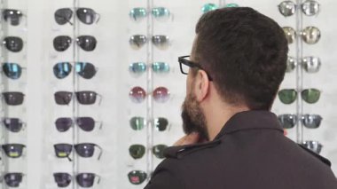 Olgun, sakallı bir adamı, bir optometrist dükkanında şık bir güneş gözlüğü dizisine bakarken yakından çeker. Düşünceli ifadesi ve canlı gözlük görüntüsü dinamik bir sahne yaratıyor.