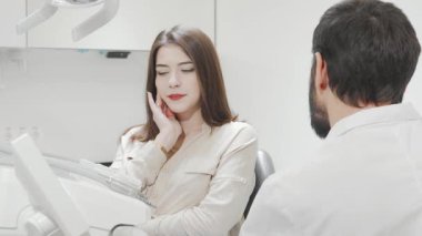 Yüzünde endişeli bir ifade olan genç bir kadın, dişçisine danışırken dişlerinin ağrıdığını belirten yanağını tutuyor. Bu sahne diş rahatsızlığının gerilimini ve parlak, steril bir şekilde sağlanan profesyonel bakımı yakalıyor.