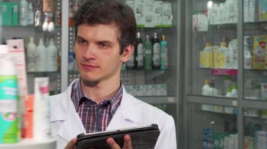 Genç, yakışıklı bir eczacı, enerjik bir eczanede envanter sayımını yaparken sıcak bir gülümsemeyle ışıldıyor. Sağlık ürünleriyle dolu renkli raflarla çevrelenmiş halde, güvenli bir şekilde dijital tablet kullanıyor, profesyonelliği somutlaştırıyor ve