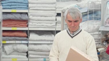 Gümüş saçlı yaşlı bir adam iyi organize edilmiş bir mobilya mağazasında renkli çarşafları incelikle tarıyor. Düzgünce istiflenmiş tekstil ürünleriyle çevrelenmiş halde, emekliliğinde rahatlık ve stil için alışverişin neşesini somutlaştırıyor.