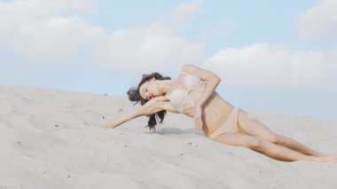 Bej bikinili çarpıcı bir kadın çölün yumuşak, sıcak kumlarında zarifçe poz veriyor, özgüven ve cazibe saçıyor. Onun zarif hareketleri ve tonlu fiziği büyüleyici bir sahne yaratıyor, şehvetin ve özgürlüğün özünü somutlaştırıyor.