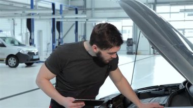 Sakallı, odaklanmış bir tamirci kaputun altındaki motoru incelerken tabletindeki araba sorunlarını özenle kaydediyor. Bu yetenekli esmer beyaz adam otomotiv sektöründe profesyonelliği somutlaştırıyor. Her detayın yakalanmasını sağlıyor.