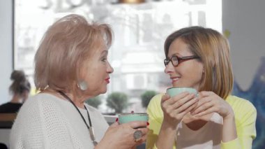 Neşeli bir kıdemli anne ve yetişkin kızı, sıcacık bir kafede renkli kahve fincanları tutarken sıcacık gülümseyerek keyifli bir anı paylaşıyorlar. Bu iç açıcı sahne, nesiller arasındaki sevgi ve bağlantının özünü yansıtıyor.