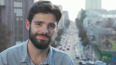 Neşeli, genç, sakallı bir adam hayat dolu hareketli bir şehir manzarasına karşı kameraya ışıldıyor. Bu yakışıklı hippi gençlik macerası ve şehir keşiflerinin özünü somutlaştırarak güven ve rahatlama yayıyor.