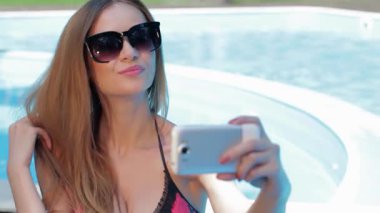Şık bikinili çarpıcı genç bir kadın, parıldayan havuzda poz verirken uzun saçları şelaleden bir selfie çeker. Neşeli bir gülümseme ve büyük güneş gözlükleriyle, neşe ve güven saçıyor, yaz eğlencesinin özünü somutlaştırıyor.