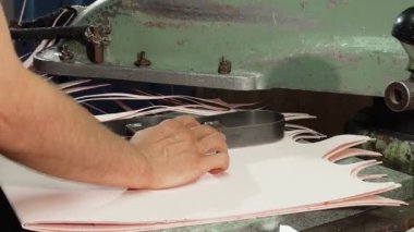 Odaklanmış bir erkek kunduracı, canlı atölyesinde titizlikle kesilmiş deri parçalarıyla çevrili bir taban kesme makinesini ustalıkla kullanır. Bu sahne, el yapımı ayakkabı üretiminin ustalığını ve hassasiyetini yansıtıyor.