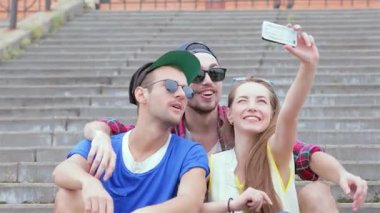 Canlı bir üçlü genç arkadaş, bir erkek ve bir kadın, mutluluk ve yoldaşlık saçan, açık hava basamaklarında selfie çekiyorlar. Parlak kıyafetleri ve şakacı ifadeleri unutulmaz bir atmosfer yaratıyor.