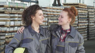 Gri iş üniforması giymiş iki neşeli genç kadın, düzgün istiflenmiş malzemelerle dolu canlı bir metal deposunda başparmaklarını gösterirken sıcak bir şekilde sarılıyorlar. Işıl ışıl gülüşleri ve yoldaşlıkları takım çalışmasının ruhunu temsil ediyor.