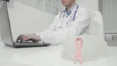 Göğüs kanseri farkındalığını sembolize eden pembe kurdeleli dokunaklı bir sahne, el değmemiş bir doktor masasında silikon göğüs implantlarıyla eşleştiriliyor. Odaklanmış ortam, kendini işine adamış bir doktorla birlikte sağlık ve önlemenin önemini vurguluyor.