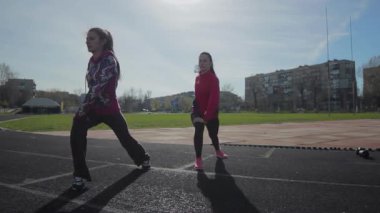 İki genç kız, yaklaşan yarışa hazırlanmak için güneş ışığında enerjik bacak kası ısınma egzersizleri yapıyorlar. Canlı spor kıyafetleri ve odaklanmış ifadeleri, bu canlı stadyum ortamında spor ve takım çalışmasına olan bağlılıklarını vurguluyor.