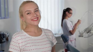 Neşeli, sarışın bir kadın, parlak bir klinikte dişlerini kontrol ettikten sonra memnuniyetle ışıldıyor. Arka planda, odaklanmış bir dişi dişçi aletlerini hazırlıyor, profesyonel ama misafirperver bir atmosfer yaratıyor.