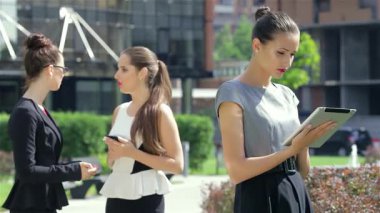 Canlı bir şehir ortamında, genç ve şık bir iş kadını tabletine sarılarak güven ve profesyonellik yayıyor. Onun kısa gülümsemesi, iki meslektaşının yakınlarda konuştuğu hareketli atmosfere biraz sıcaklık katıyor.