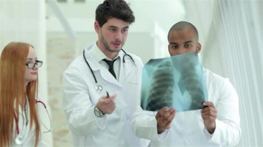 Modern bir hastane ortamında, kızıl saçlı ve gözlüklü bir kadın da dahil olmak üzere üç kendine güvenen doktor, akciğer sağlığının önemini vurgulayarak sağlıklı akciğerlerin röntgenini analiz ediyor. Odaklanmış ifadeleri ve profesyonel kıyafetleri, takım çalışmalarını ve...