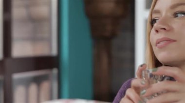 Genç, çekici, sarışın bir kadının yakın plan fotoğrafı. Çenesini eline dayamış, hareketli bir kafede düşüncelere dalmış. Ferahlatıcı bir içecek tutuyor, pencereden dışarı bakıyor, hareketli şehrin ortasında huzurlu bir anı somutlaştırıyor.