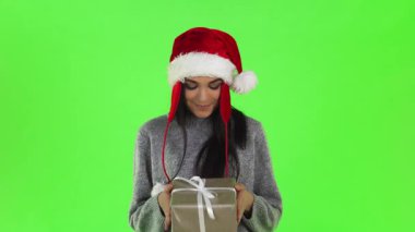 Parlak yeşil bir geçmişi olan canlı bir stüdyo ortamında, Noel Baba gibi giyinmiş neşeli genç bir kadın güzel paketlenmiş bir Noel hediyesini elinde tutuyor. Neşeli ruhu sıcaklık ve heyecan saçıyor, herkesi paylaşmaya davet ediyor.