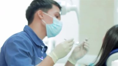 Maske ve eldiven takan odaklanmış genç bir dişçi hastaların dişlerini hassas aletlerle dikkatlice inceler. Parlak klinik ayarı özel bir lambayla aydınlatılır. Titiz teftiş sürecini geliştirir ve en uygun diş bakımını sağlar.