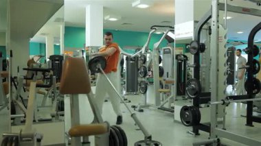 Orta Doğulu etnik kökenli odaklanmış genç bir adam canlı bir spor salonunda halteri tutkulu bir şekilde kaldırıyor ve göğüs kasları ve pazılarını geliştirmedeki kararlılığını gösteriyor. Çeşitli fitness ekipmanlarıyla dolu, parlak, modern spor salonu,