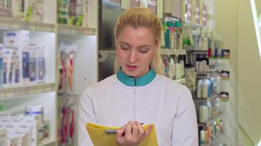 Kıtır kıtır laboratuvar önlüğü giymiş neşeli genç bir kadın eczacı etrafı düzenli ilaç raflarıyla çevrili iken dikkatle evrak dolduruyor. Onun odaklanmış ifadesi ve canlı sarı saçı onun mükemmel olmaya olan bağlılığını yansıtıyor.