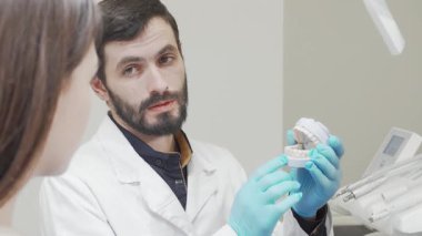 İyi bakımlı sakallı odaklanmış bir erkek dişçi modern bir klinikteki meraklı hastasına detaylı bir diş kalıbı gösteriyor. Parlak, steril çevre diş sağlığının önemini vurgularken, diş hekimlerinin arkadaş canlısı tavırları diş sağlığını teşvik eder.