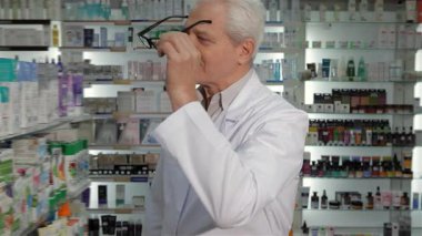 Gözlüklü ve beyaz önlüklü seçkin bir erkek eczacı, düşünceli bir şekilde dolu bir eczanede duruyor. Gri saçları ve sakin tavırları, sağlık ürünlerini dikkatlice incelerken yıllar süren deneyimini yansıtıyor.
